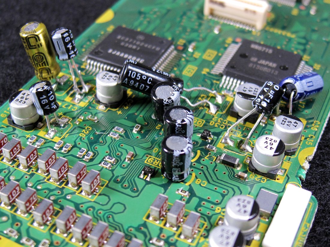 Six mismatched through-hole capacitors standing on bent leads over surface-mount pads on an AG-1980 timebase corrector board, one lying on its side, beside the untouched original parts